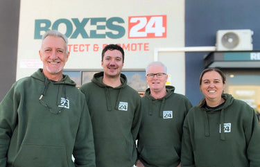 Four people wearing green hoodies with a 'Boxes24' logo in front of a 'Boxes24' branded vehicle.
