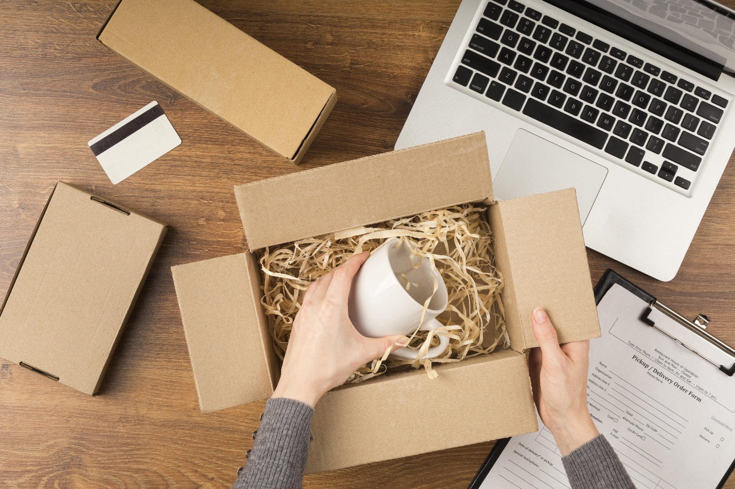 Person packing a white mug into a cardboard box on a wooden table with a laptop and other items.