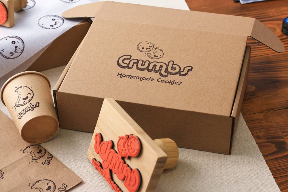 Crumbs Homemade Cookies packaging with a box, cup, and cookie stamp on a wooden surface.